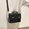 A black minimalist leather crossbody bag is shown hanging from a strap against a white shirt.