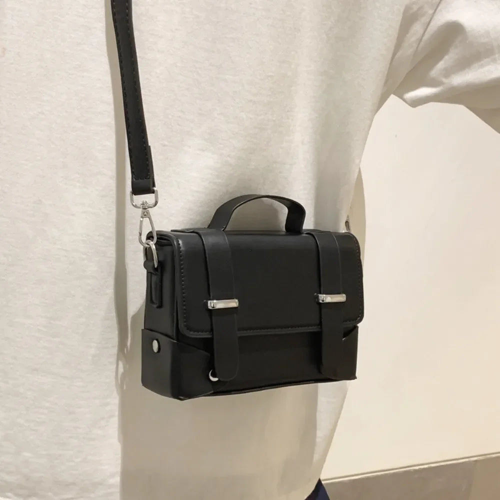 A black minimalist leather crossbody bag is shown hanging from a strap against a white shirt.