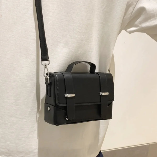 A black minimalist leather crossbody bag is shown hanging from a strap against a white shirt.