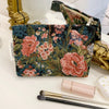 A floral patterned velvet cosmetic pouch next to makeup brushes and gold jewelry.