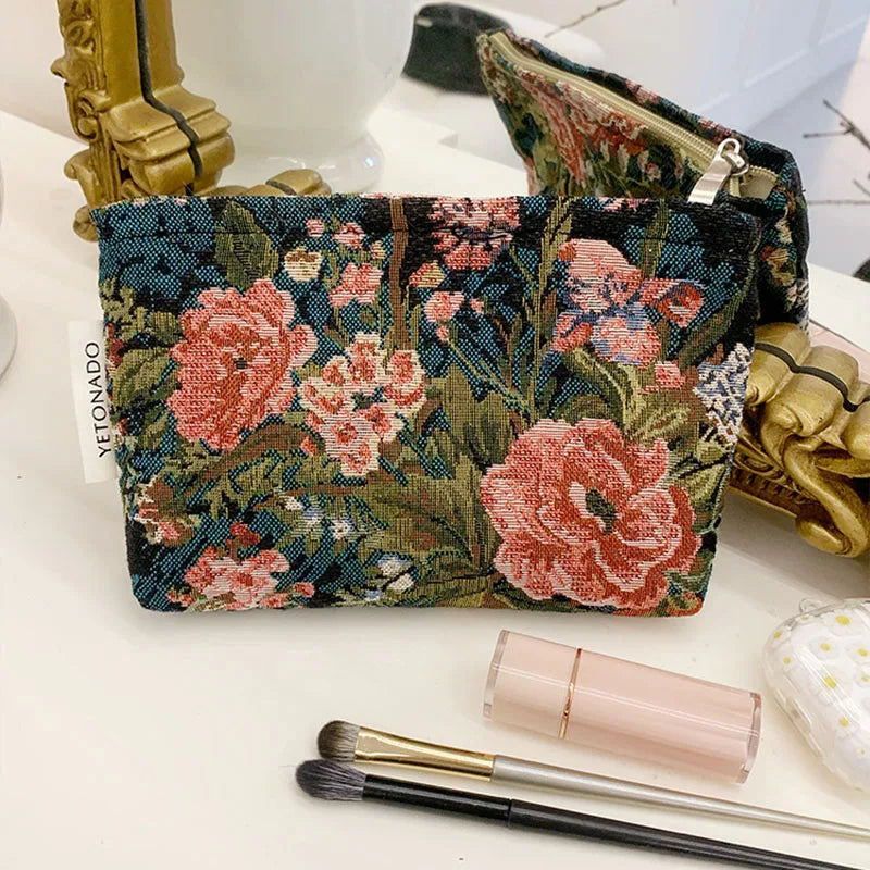 A floral patterned velvet cosmetic pouch next to makeup brushes and gold jewelry.