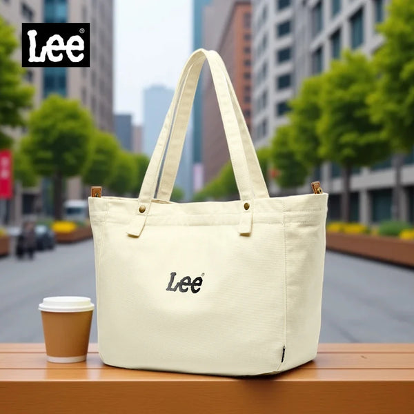 Un sac fourre-tout Lee en toile Oxford de couleur crème est exposé sur une surface en bois dans un cadre urbain extérieur.