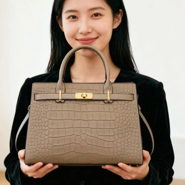 Eine Frau hält eine beige Lederhandtasche mit goldfarbenen Verzierungen vor weißem Hintergrund.