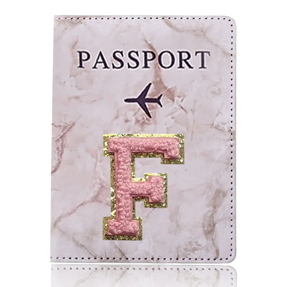 Una funda de pasaporte de imitación de cuero rosa claro con una inicial F dorada y un ícono de avión negro.