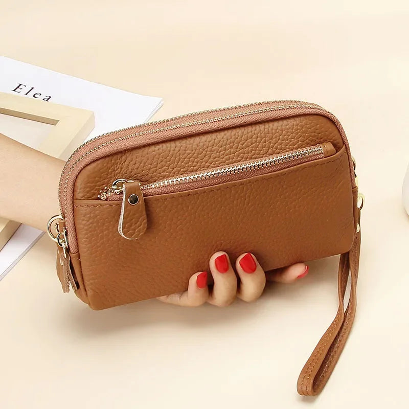 A brown genuine cowhide leather double-zip wristlet wallet is held in a hand on a light background.