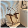 A beige canvas shoulder tote bag with tan leather accents resting on a dark surface next to a window.