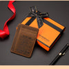 Une boîte cadeau orange attachée d'un ruban noir, à côté d'un portefeuille RFID en cuir marron et d'un stylo noir.