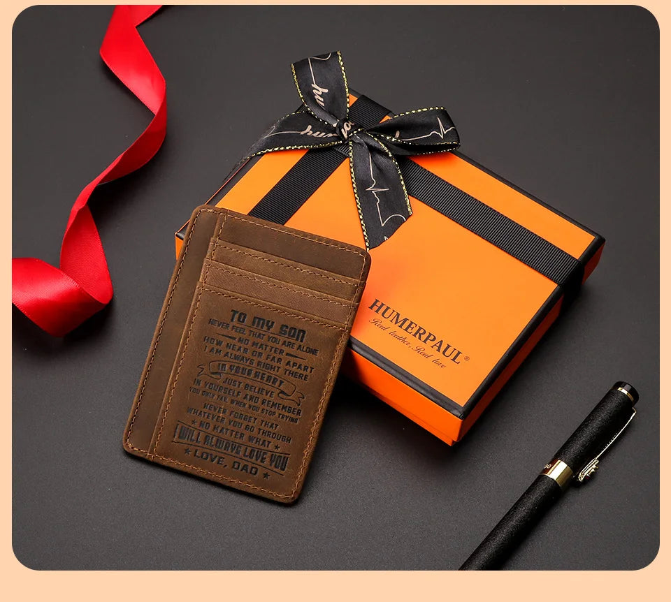 Une boîte cadeau orange attachée d'un ruban noir, à côté d'un portefeuille RFID en cuir marron et d'un stylo noir.