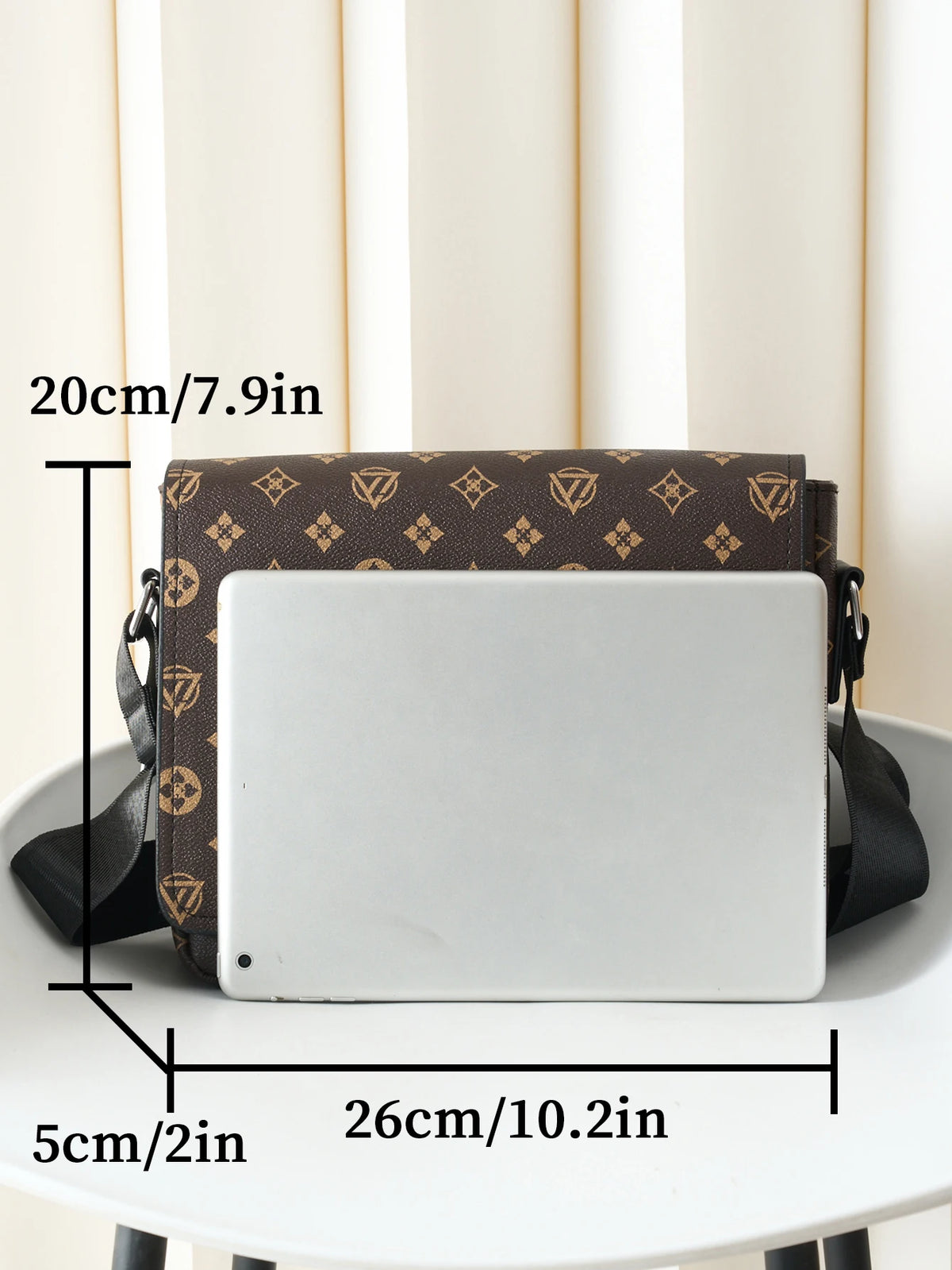 A brown leather crossbody bag with a flap design and a silver-colored laptop inside, shown on a white table.