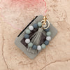 A light gray leather card holder with a beaded tassel keychain in muted blue and white tones.