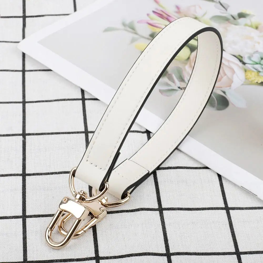 A beige leather replacement shoulder strap with a gold-toned clasp is displayed on a white and black checkered surface.