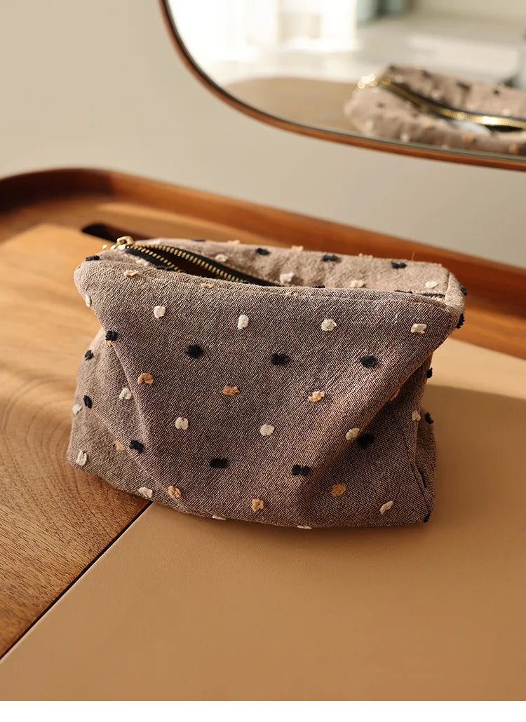 Pochette de rangement cosmétique en tissu gris plié avec de petits pois noirs et dorés sur une surface en bois.