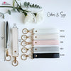 A collection of leather wristlet keychains in various colors including white, pink, grey, and black, displayed with greenery.