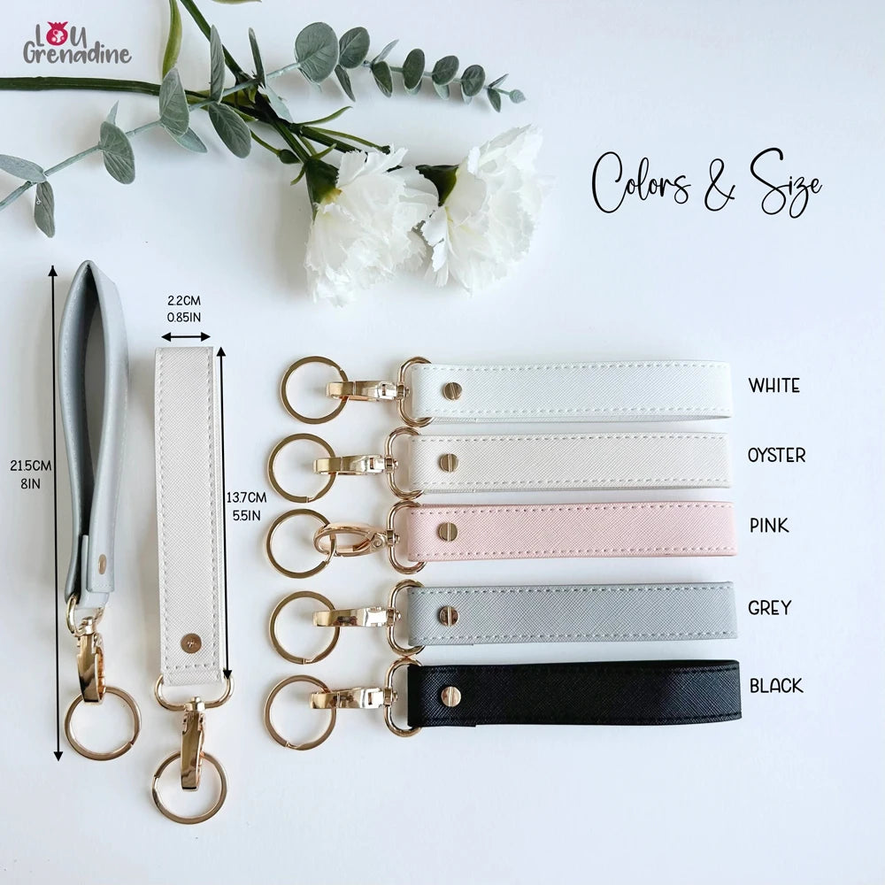 A collection of leather wristlet keychains in various colors including white, pink, grey, and black, displayed with greenery.
