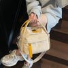 A light beige nylon crossbody bag with a yellow top handle is being held by a person's hand while standing on dark wooden sta