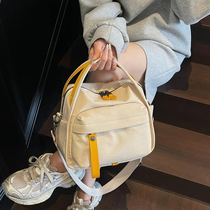 A light beige nylon crossbody bag with a yellow top handle is being held by a person's hand while standing on dark wooden sta