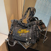 A dark gray denim crossbody bag with gold hardware and yellow embroidery is displayed on a dark surface.
