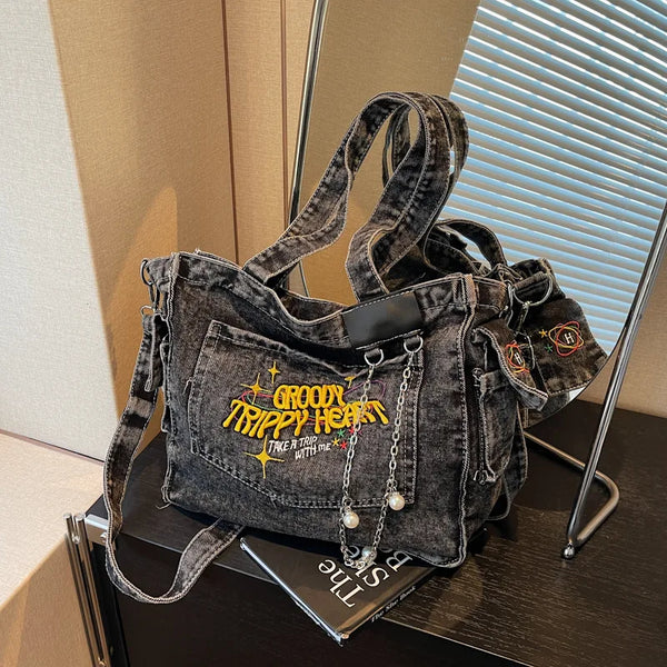A dark gray denim crossbody bag with gold hardware and yellow embroidery is displayed on a dark surface.