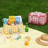 A red and white checkered insulated picnic tote bag filled with snacks and a yellow checkered tablecloth on green grass.