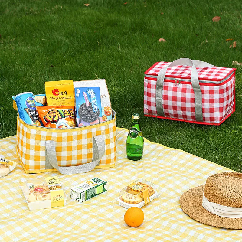 A red and white checkered insulated picnic tote bag filled with snacks and a yellow checkered tablecloth on green grass.