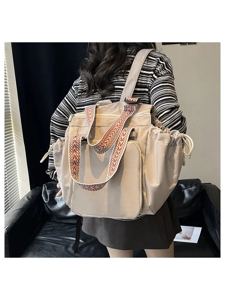 A woman is holding a beige nylon shoulder bag with striped leather straps in a casual indoor setting.