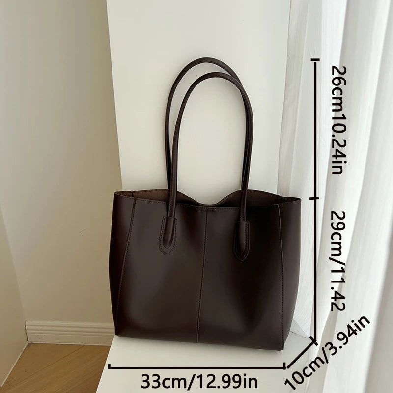 A dark brown leather tote bag hanging against a white wall with a measurement scale.