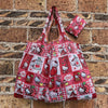 A pink and red checkered reusable shopping bag featuring cute cat designs on a brick wall.