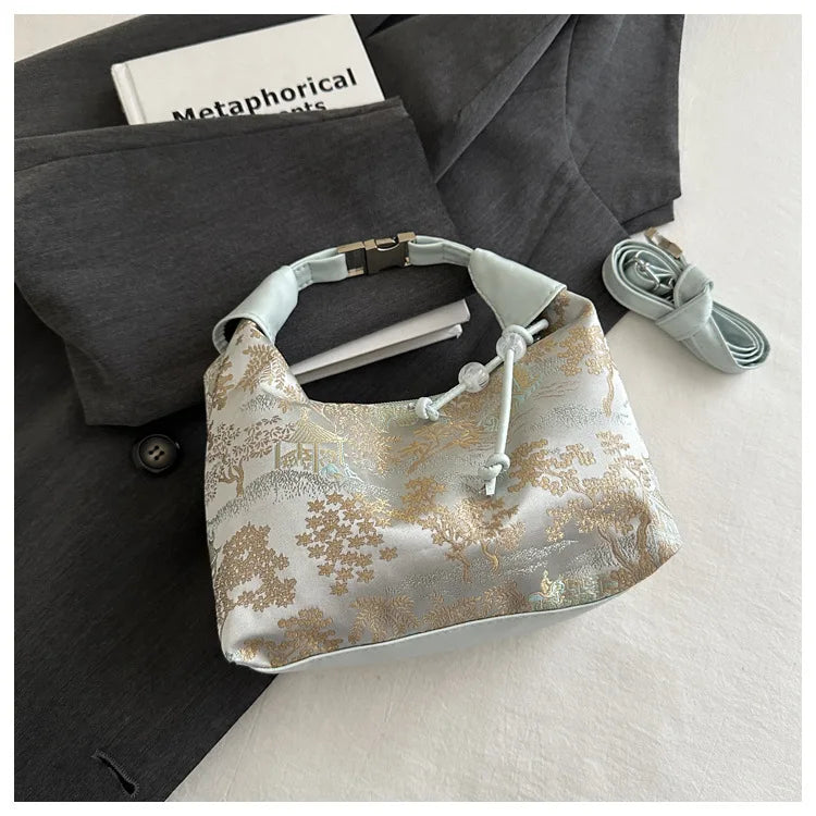 A light beige embroidered crossbody shoulder bag resting on a dark gray blazer with a white pen.