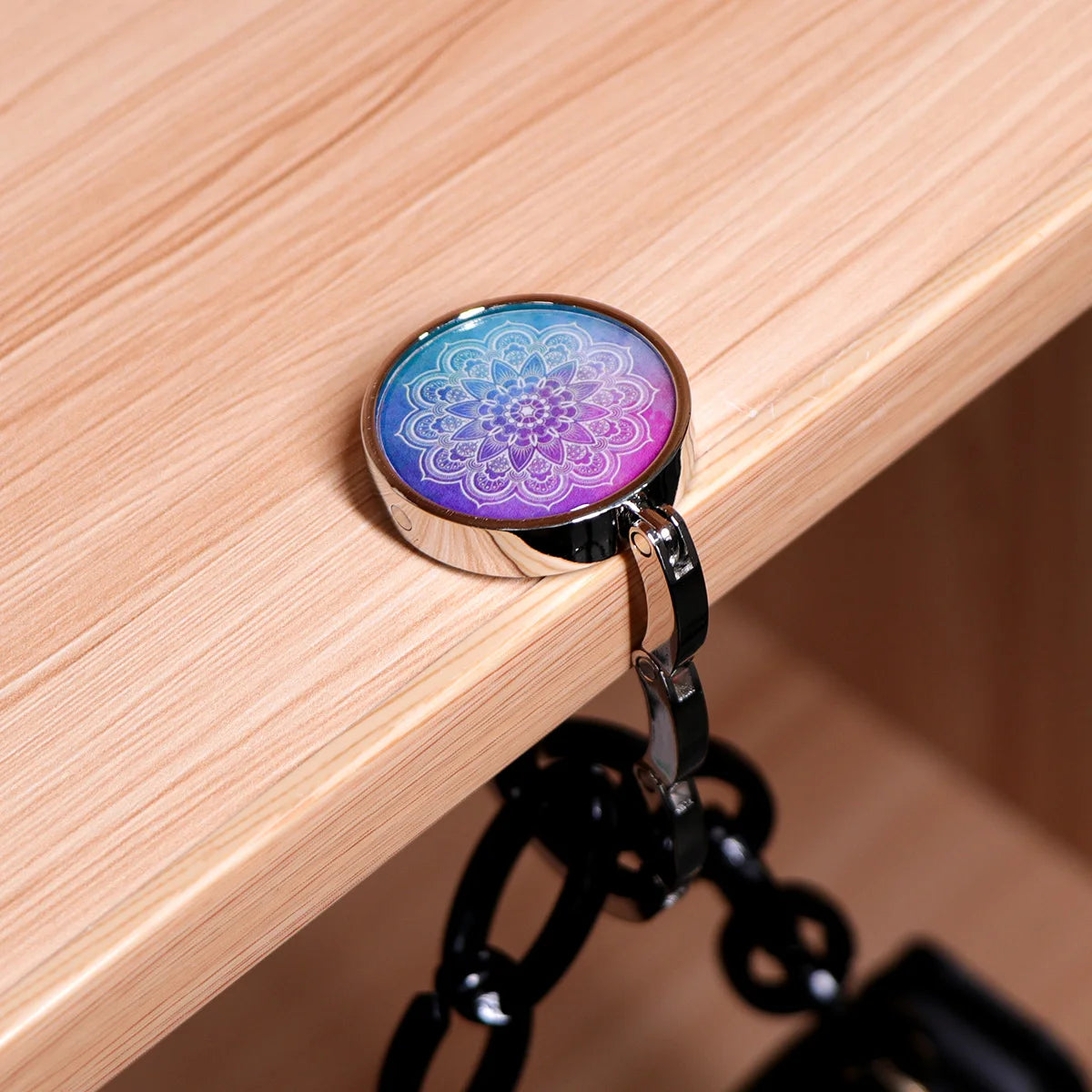 A silver folding table bag hook with a round, iridescent marble design is shown resting on a light wood surface.