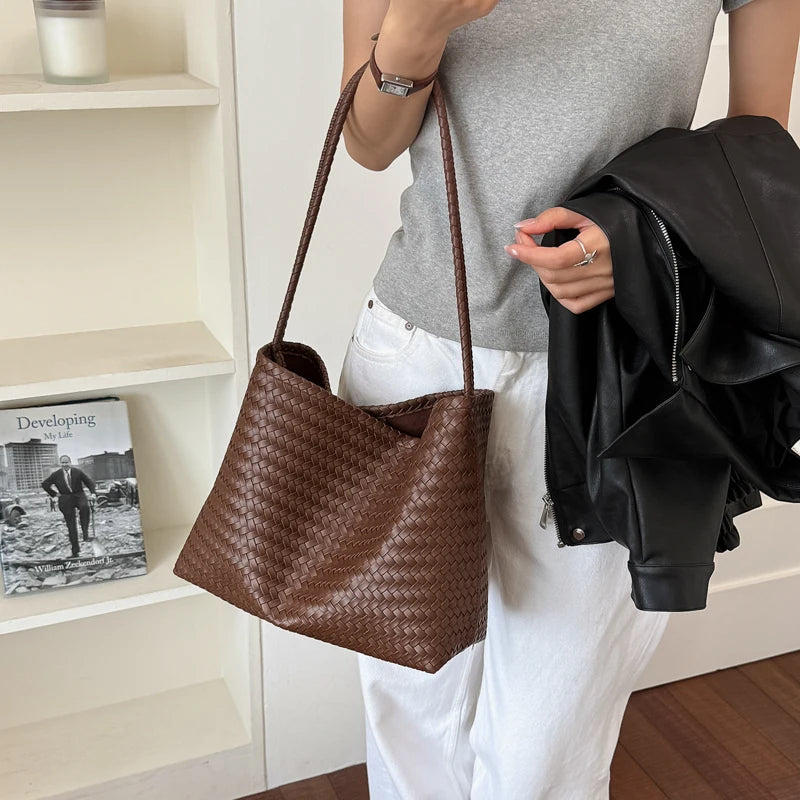 A woman holds a brown woven PU bucket shoulder bag and a black leather jacket in a bright indoor setting.