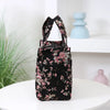 A black cloth tote bag with a pink floral pattern is displayed on a white surface next to light blue decorative objects.