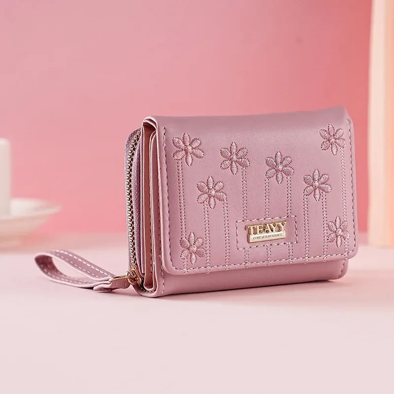 A light pink compact PU leather women's wallet with a zipper and floral embroidery is displayed on a pink surface.