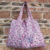 A pink reusable shopping bag featuring a pattern of cute cats against a white background, displayed against a brick wall.