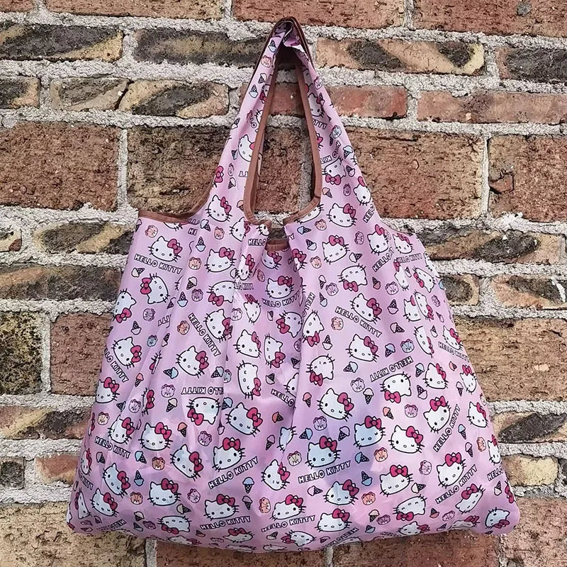A pink reusable shopping bag featuring a pattern of cute cats against a white background, displayed against a brick wall.