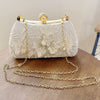 A white clutch bag with gold chain straps and a pearl embellishment rests on a wooden surface.