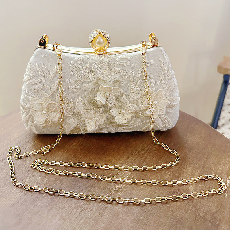 A white clutch bag with gold chain straps and a pearl embellishment rests on a wooden surface.