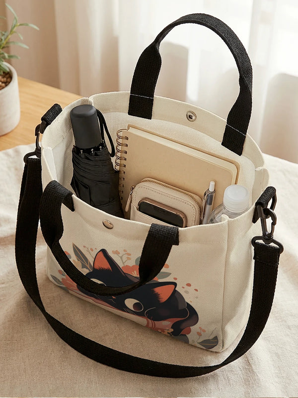 A cream-colored tote bag with a floral cat print and a black strap, holding a pen and small items.