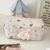 A pink cosmetic storage pouch with white quilted fabric and a floral pattern rests next to a light-colored decorative item.