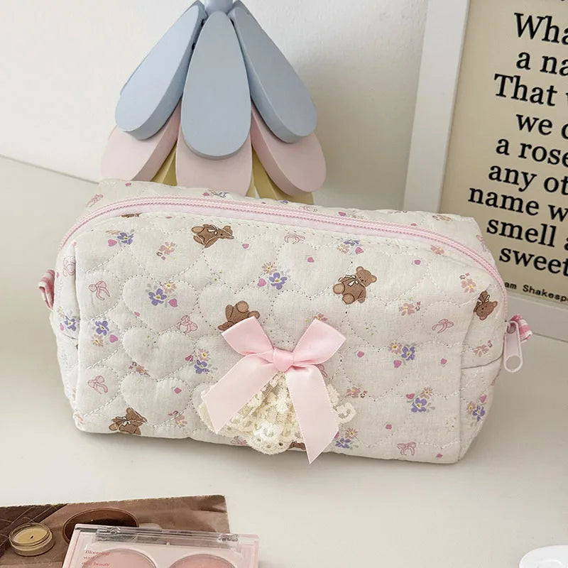A pink cosmetic storage pouch with white quilted fabric and a floral pattern rests next to a light-colored decorative item.