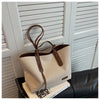 A beige canvas shoulder tote bag with brown leather accents is displayed in front of white slatted shutters.
