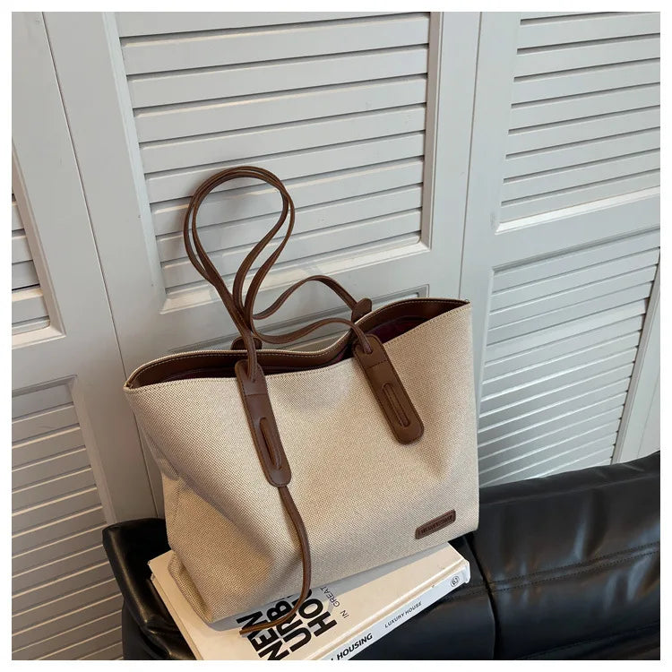 A beige canvas shoulder tote bag with brown leather accents is displayed in front of white slatted shutters.