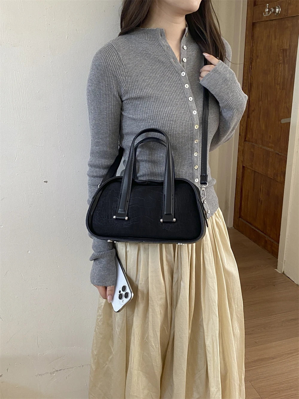 A woman wearing a grey knitted top and a cream skirt holds a black crocodile textured mini crossbody bag.