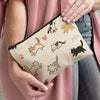 A beige cosmetic pouch featuring cartoon cats in various poses, held by a person wearing a pink cardigan.