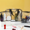A set of three clear PVC travel toiletry bags with black straps and various toiletries displayed on a white surface.