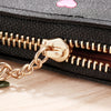 A black compact PU leather wallet with a gold-toned chain closure and zipper is shown on a wooden surface.