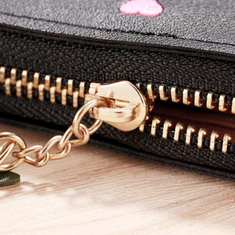 A black compact PU leather wallet with a gold-toned chain closure and zipper is shown on a wooden surface.