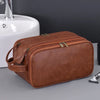 A brown PU leather travel toiletry bag with organizer pockets resting on a gray surface next to a white sink.