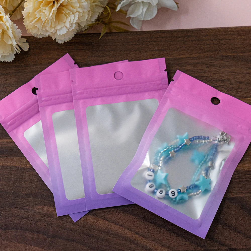 Three resealable gradient storage bags in pink and purple holding jewelry or supplies on a wooden surface.