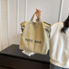 A light beige canvas tote bag is displayed on a dark surface next to a white sweater.