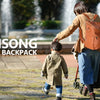 A woman wearing a brown jacket and blue pants carries a Minsong diaper backpack while pushing a stroller in a park.